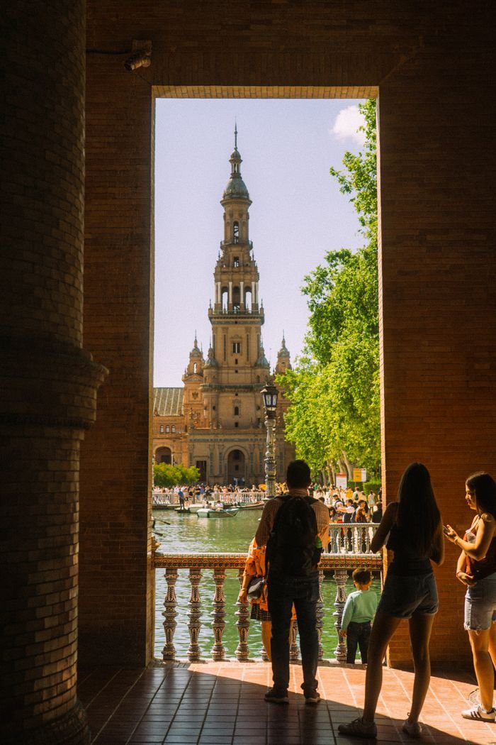 Cookie Policy View of Plaza de España in Seville through an archway with people silhouetted, capturing historic architecture.